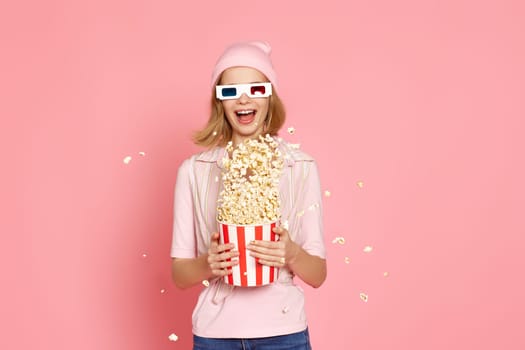 Cheerful blonde woman in 3d-glasses holding bucket of popcorn and jumping on pastel pink background.