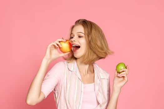 beautiful blonde woman holding green and yellow apples on pink background. healthy food