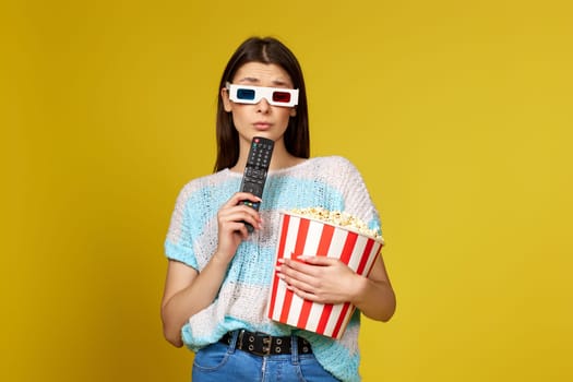 thoughtful brunette woman with remote control watching interesting TV series and holding bucket of popcorn on yellow background.