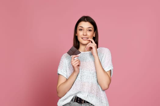 happy smiling girl in sweater holding delicious chocolate bar on pink background