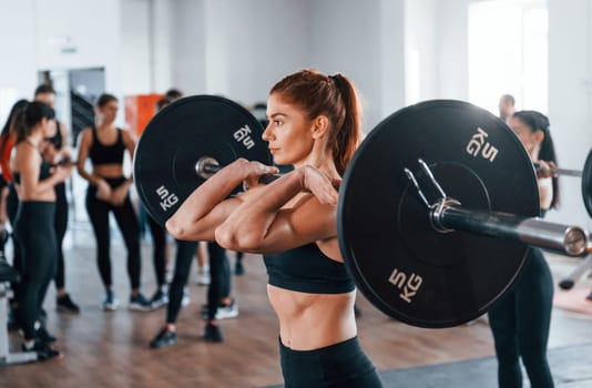 Squats with barbell. Group of young sportive people have crossfit day indoors in gym.