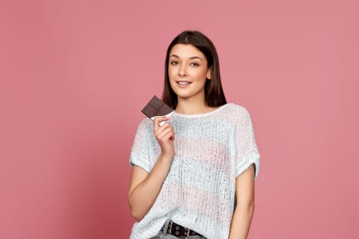 happy smiling girl in sweater holding delicious chocolate bar on pink background