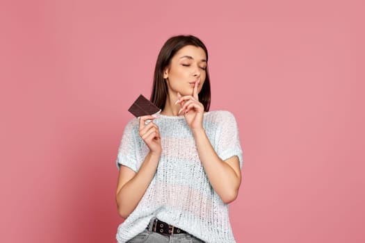 young woman in sweater holding delicious chocolate bar shows a gesture quietly on pastel pink background