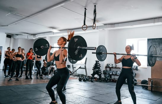 Squats with barbell. Group of young sportive people have crossfit day indoors in gym.