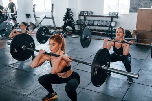 Squats with barbell. Group of young sportive people have crossfit day indoors in gym.