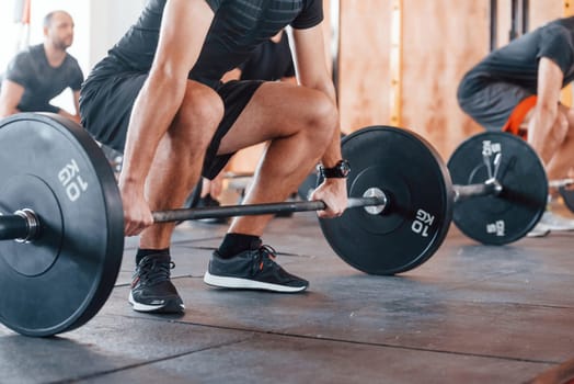 By using heavy weights. Group of young sportive people have crossfit day indoors in gym.