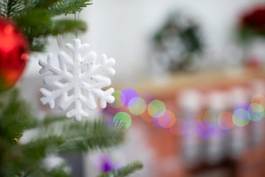 Snowflake-shaped bauble hangs on the branch of the Christmas tree. Fireplace in backgourd.