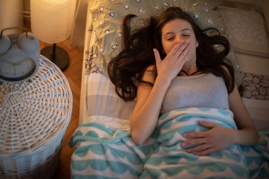 A yawning woman lies in her bed with the bedside lamp on. A girl under a soft blanket.