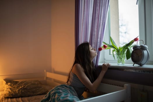 A young woman sniffs the tulips in a vase on the windowsill. Bright bedroom with decorations.