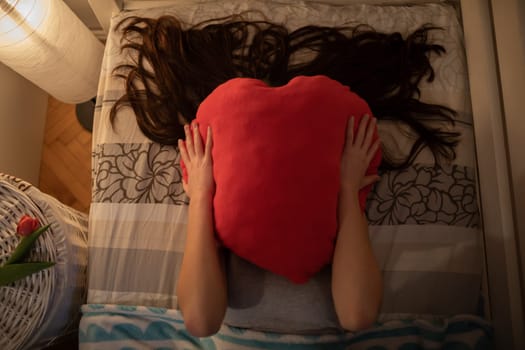 The girl covers her face with a red heart-shaped pillow. The light of a lamp on the wall.