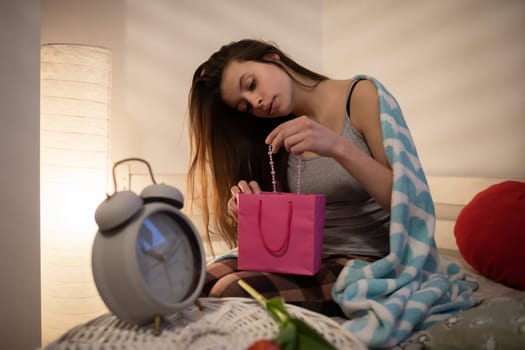 Valentine's Day gift on girl's lap wrapped in pink bag. Bright bedroom with decorations.