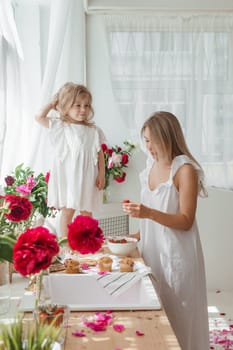 A little blonde girl with her mom on a kitchen countertop decorated with peonies. The concept of the relationship between mother and daughter. Spring atmosphere