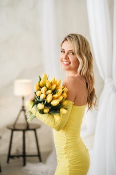 a happy woman in a yellow dress embraces a bouquet of yellow spring tulips in the interior.