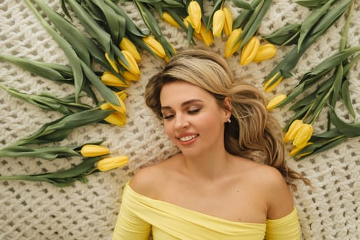 a happy woman in a yellow dress lies in yellow spring tulips on the bed.