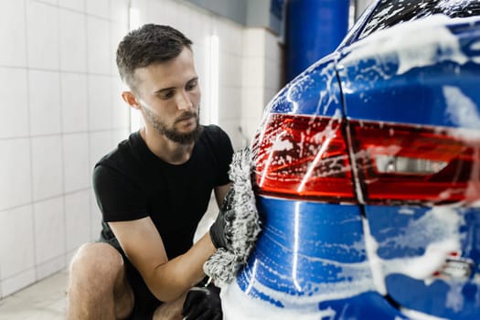 Car washer doing manual foam washing in auto detailing service. Hand washing with microfiber glove with foam car body in garage