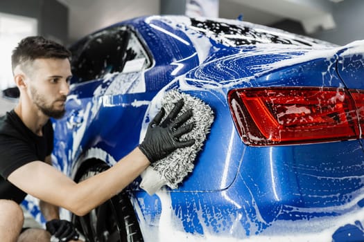 Car washer doing manual foam washing in auto detailing service. Hand washing with microfiber glove with foam car body in garage