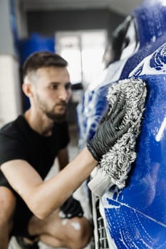 Car washer doing manual foam washing in auto detailing service. Hand washing with microfiber glove with foam car body in garage