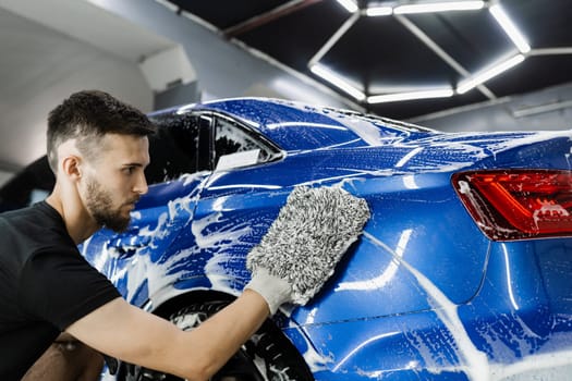 Car washer doing manual foam washing in auto detailing service. Hand washing with microfiber glove with foam car body in garage