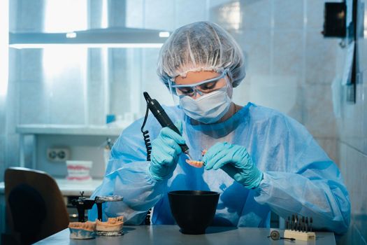 A masked and gloved dental technician works on a prosthetic tooth in his lab.