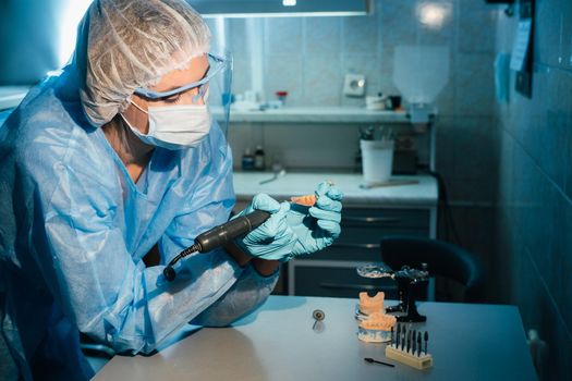 A masked and gloved dental technician works on a prosthetic tooth in his lab.