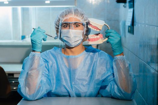 A dental doctor wearing blue gloves and a mask holds a dental model of the upper and lower jaws and a dental mirror.