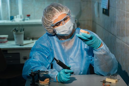 A masked and gloved dental technician works on a prosthetic tooth in his lab.