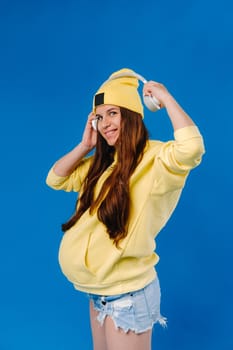 A pregnant girl in a yellow jacket and headphones stands on a blue background.