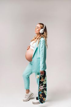 A pregnant girl in a turquoise suit with a skateboard in her hands and headphones stands on a gray background.