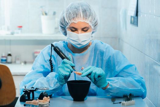 A masked and gloved dental technician works on a prosthetic tooth in his lab.