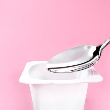 Yogurt cup and silver spoon on pink background, white plastic container with yoghurt cream, fresh dairy product for healthy diet and nutrition balance.