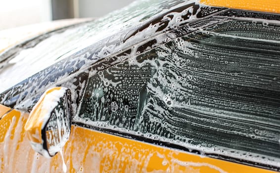 Side window and mirror of yellow car washed in carwash. Strokes from brush visible on glass in white shampoo and foam.