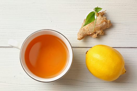 Tabletop view, porcelain cup of freshly brewed tea, dry ginger root with green sprout and whole lemon next to it, placed on white boards desk.