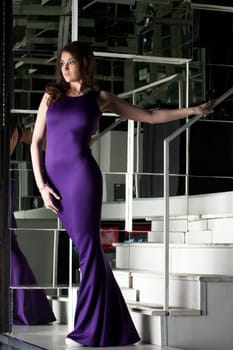 Full length portrait of pretty brunette woman in long purple dress posing on stairs