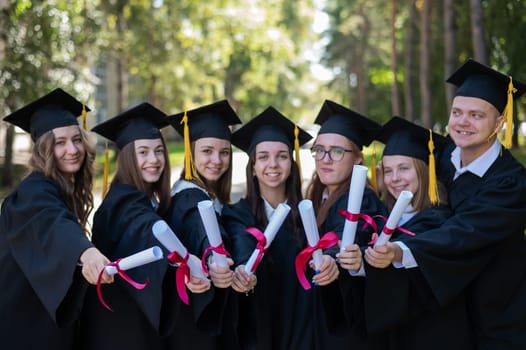 Row of happy young people in graduation gowns holding diplomas outdoors