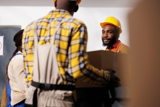 Warehouse manager looking at colleague bringing parcel to reception desk in storage room. African american storehouse employee wearing protective hard hat receiving box from package handler