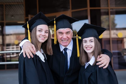 An elderly man hugs young girls. Classmates in graduation gowns