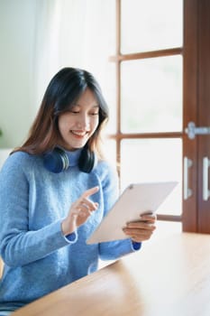 Portrait of a teenage Asian woman using a tablet, wearing headphones to study online via video conferencing on a wooden desk in library.