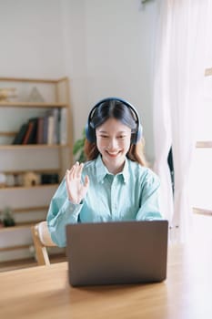 Portrait of a teenage Asian woman using a computer, wearing headphones to study online via video conferencing on a wooden desk at home.