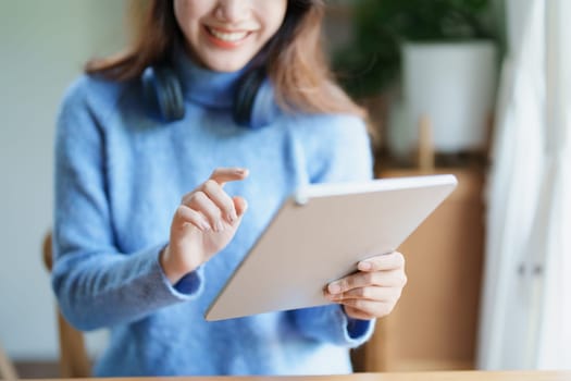 Portrait of a teenage Asian woman using a tablet, wearing headphones to study online via video conferencing on a wooden desk in library.