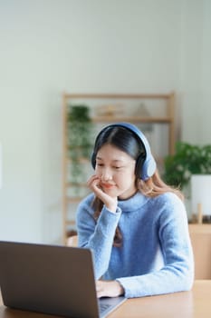 Portrait of a teenage Asian woman using a computer, wearing headphones to study online via video conferencing on a wooden desk at home.