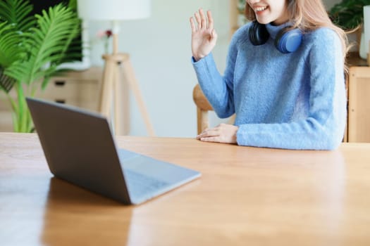 Portrait of a teenage Asian woman using a computer, wearing headphones to study online via video conferencing on a wooden desk at home.