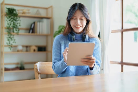 Portrait of a teenage Asian woman using a tablet, wearing headphones to study online via video conferencing on a wooden desk in library.