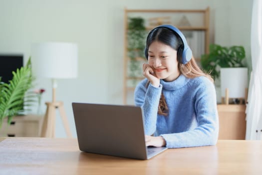 Portrait of a teenage Asian woman using a computer, wearing headphones to study online via video conferencing on a wooden desk at home.