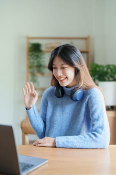 Portrait of a teenage Asian woman using a computer, wearing headphones to study online via video conferencing on a wooden desk at home.