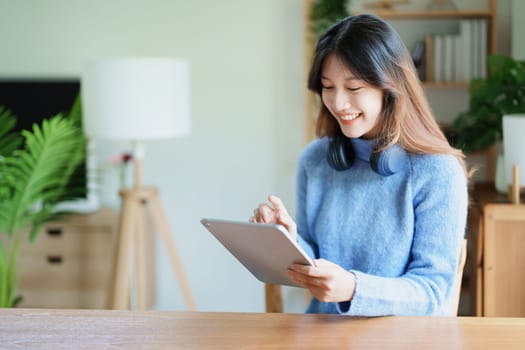 Portrait of a teenage Asian woman using a tablet, wearing headphones to study online via video conferencing on a wooden desk in library.