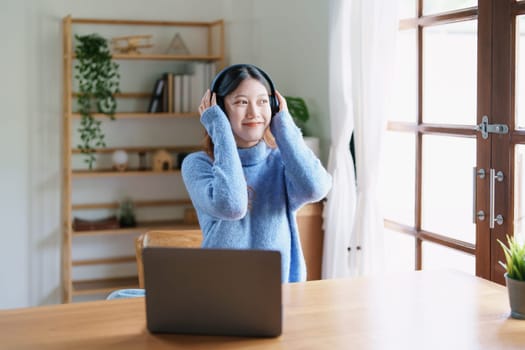 Portrait of a teenage Asian woman using a computer, wearing headphones to study online via video conferencing on a wooden desk at home.