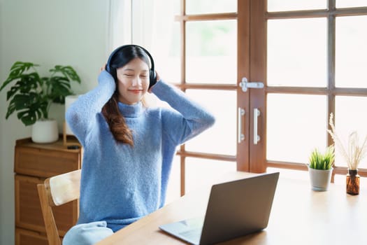 Portrait of a teenage Asian woman using a computer, wearing headphones to study online via video conferencing on a wooden desk at home.