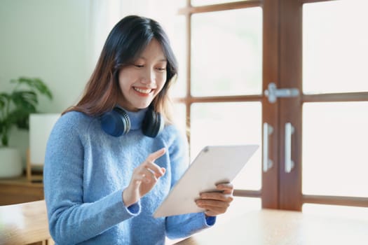 Portrait of a teenage Asian woman using a tablet, wearing headphones to study online via video conferencing on a wooden desk in library.