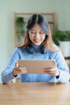 Portrait of a teenage Asian woman using a tablet, wearing headphones to study online via video conferencing on a wooden desk in library.