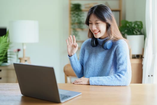 Portrait of a teenage Asian woman using a computer, wearing headphones to study online via video conferencing on a wooden desk at home.